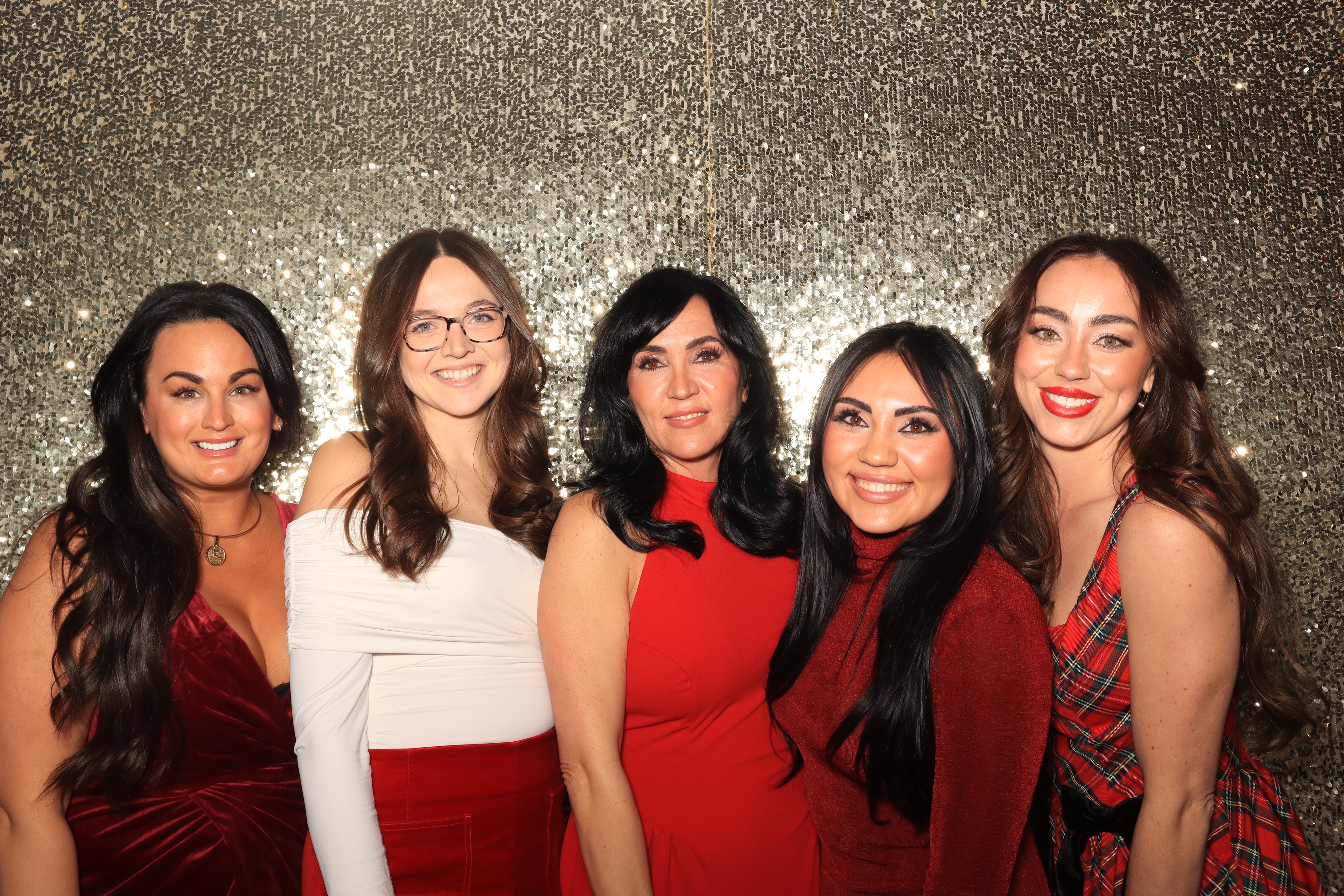 Five guests in red dresses smiling together in front of a gold sequin backdrop at the Festival of Trees photo booth